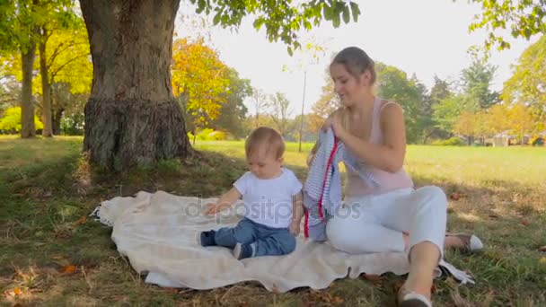 Jovem mãe sentada com seu bebê na sombra sob a árvore e vestir o filho no casaco — Vídeo de Stock