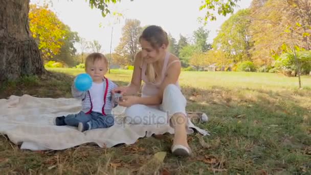 Feliz joven madre sentada con su hijo bebé en la manta bajo el árbol en el parque — Vídeos de Stock