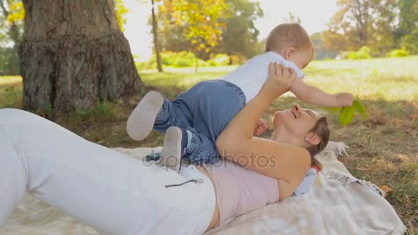 Jovem mãe feliz brincando em cobertor com seu filho bebê debaixo da árvore no parque — Vídeo de Stock