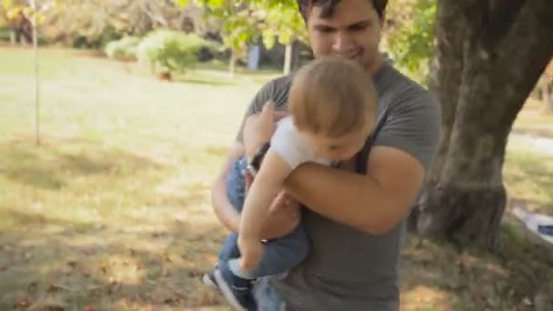 公園の木の下で彼の赤ん坊の息子を抱きしめる幸せな若い父の肖像 — ストック動画