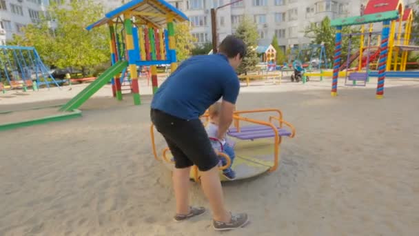 Счастливый молодой отец катается на своем сыне на карусели на детской площадке — стоковое видео