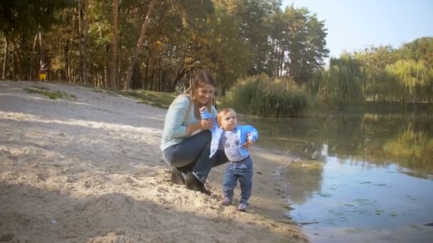 美丽年轻的母亲带着她的宝贝儿子坐在沙滩上，看着美丽的湖，在森林里 — 图库视频影像