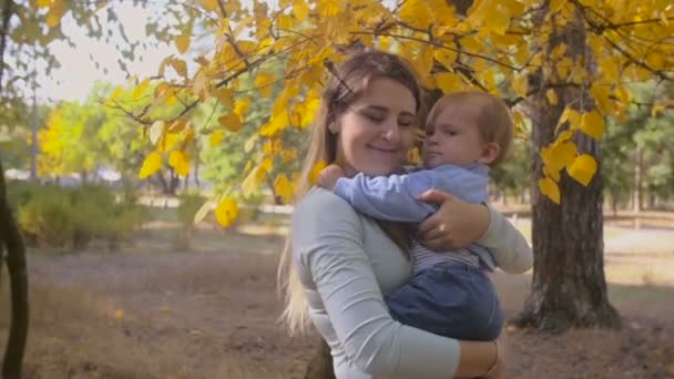 Filmagem em câmera lenta da jovem mãe segurando e abraçando seu filho no parque de outono — Vídeo de Stock