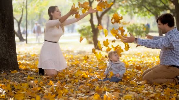 Повільні кадри руху щасливої молодої сім'ї, що кидає листя з дитиною на осінній парк — стокове відео