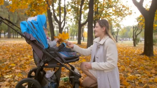 Glückliche junge Mutter schenkt ihrem Baby im Kinderwagen im Herbstpark gelbe Blätter — Stockvideo