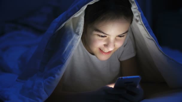 Портрет усміхненої дівчини-підлітка, що лежить під ковдрою і використовує мобільний телефон — стокове відео