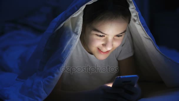 Retrato de una adolescente sonriente acostada debajo de una manta y usando un teléfono móvil — Vídeos de Stock