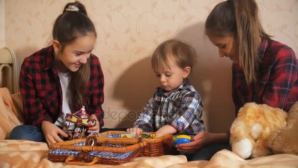 Милый мальчик играет в игрушки со своими сестрами на кровати в спальне — стоковое видео