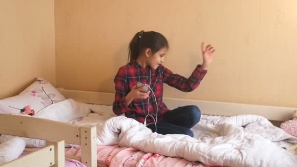 Vídeo em câmera lenta de uma adolescente alegre sentada na cama e ouvindo música com telefone celular — Vídeo de Stock