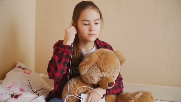 Slow motion beelden van tienermeisje luisteren naar muziek met een koptelefoon en teddy bear knuffelen — Stockvideo