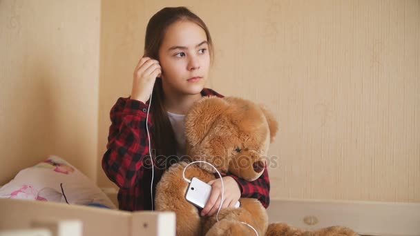 Slow motion footage of upset teenage girl listening to music and holding teddy bear on bed — Stock Video