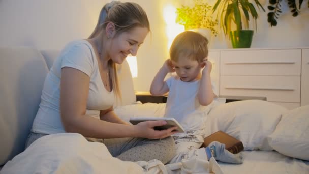 4k видео молодой матери с малышом, играющей на планшете в постели — стоковое видео