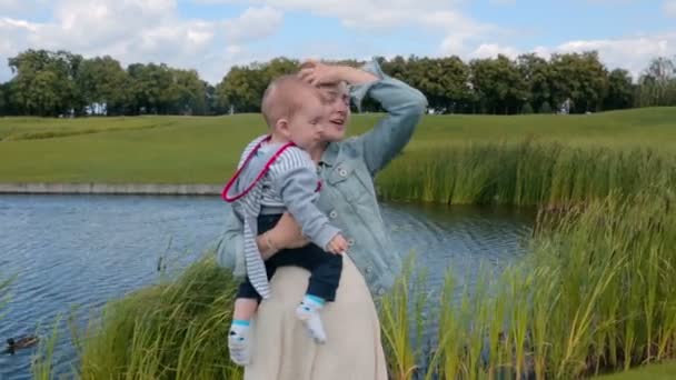 快乐年轻的母亲抱着她 1 岁的儿子在公园的大池塘 — 图库视频影像