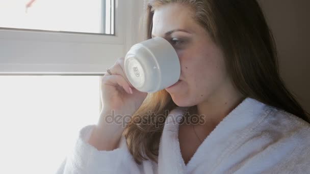 Видеозапись молодой женщины в халате, пьющей кофе и смотрящей в окно — стоковое видео
