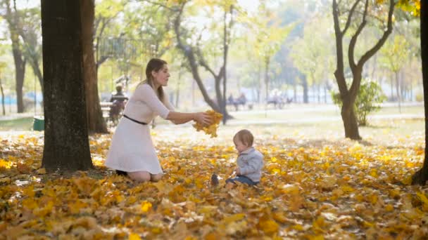 Movimiento lento de la madre feliz con el bebé sentado en el parque y lanzando hojas — Vídeos de Stock