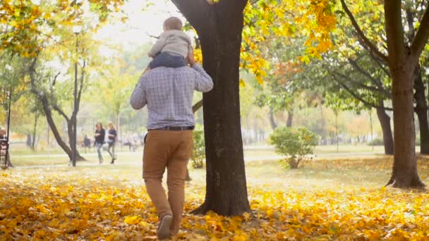 Images au ralenti de bébé mignon assis sur les épaules des pères marchant au parc d'automne — Video