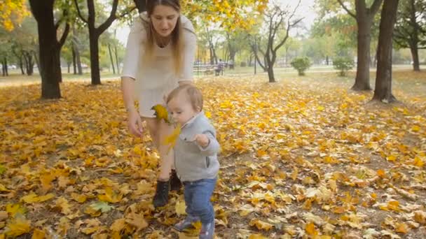 Images au ralenti de mignon bébé garçon tenant des feuilles d'automne et marchant avec sa mère au parc 