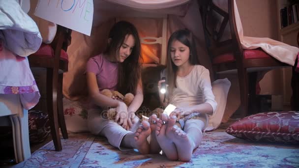 4 k βίντεο των δύο χαριτωμένα κορίτσια διαβάζοντας το βιβλίο με το φακό στο σπίτι κατασκευασμένο από κουβέρτες — Αρχείο Βίντεο