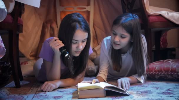 4k video of two sisters lying in house made of blankets and reading book with torch — Stock Video