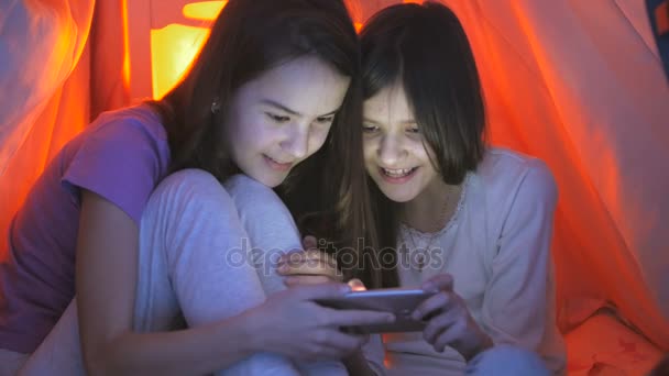 Due ragazze sorridenti in pigiama che giocano sullo smartphone di notte sotto coperta — Video Stock