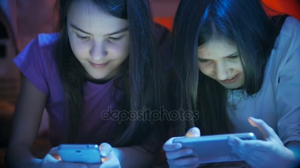 Съемки крупным планом двух симпатичных девушек, лежащих в спальне и использующих мобильные телефоны поздно ночью — стоковое видео