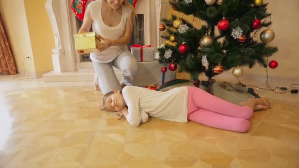 Χαριτωμένο κορίτσι αποκοιμήθηκε κάτω από το χριστουγεννιάτικο δέντρο. Μητέρα της εγρήγορσης και δίνοντας το δώρο Χριστουγέννων — Αρχείο Βίντεο