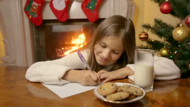 Porträtt av söt leende flicka sitter bakom bordet i vardagsrummet och skriva brev till jultomten — Stockvideo
