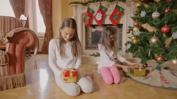 Dos chicas alegres en pijama sentadas bajo el árbol de Navidad y abriendo cajas con regalos — Vídeos de Stock