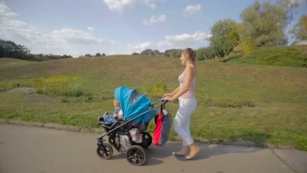 Hermosa mujer sonriente caminando con hijo de la bahía en cochecito en el parque — Vídeo de stock