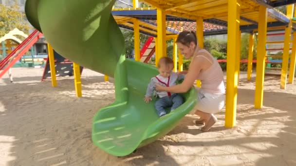 Lindo niño sonriente cabalgando en las diapositivas con la madre en el patio de recreo — Vídeo de stock