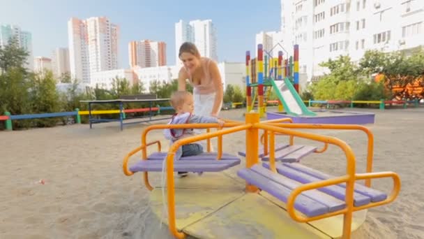 Gelukkig jonge moeder die haar zoontje op de carrousel op speelplaats spinnen — Stockvideo