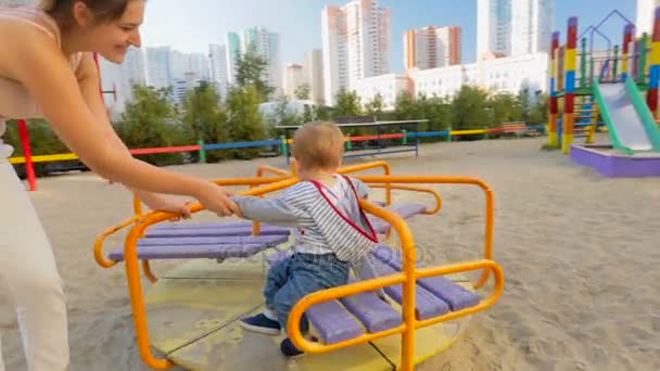 Feliz madre sonriente montando a su hijo bebé en carrusel en el patio de recreo — Vídeo de stock