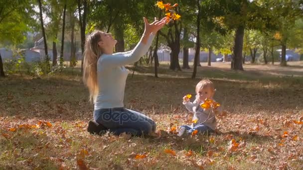 Jovem mãe feliz com seu filho bebê brincando com folhas caídas no parque — Vídeo de Stock