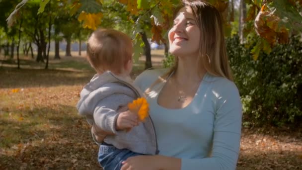 Slow motion beelden van gelukkige jonge moeder die haar zoontje onder boom in herfst park knuffelen — Stockvideo