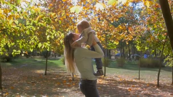 Filmagem em câmera lenta da bela jovem mãe segurando e levantando no ar seu filho bebê no parque de outono — Vídeo de Stock