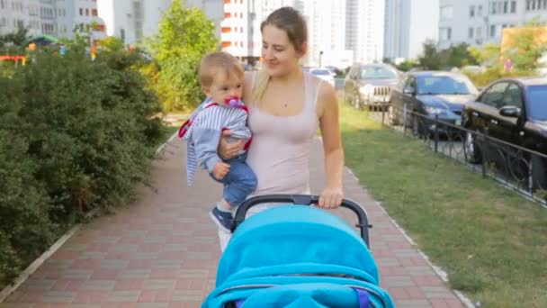 Щаслива усміхнена мати тримає свого сина і штовхає коляску на вулицю — стокове відео