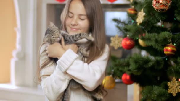 Retrato de linda chica alegre acariciando su lindo gatito gris junto al árbol de Navidad decorado — Vídeos de Stock