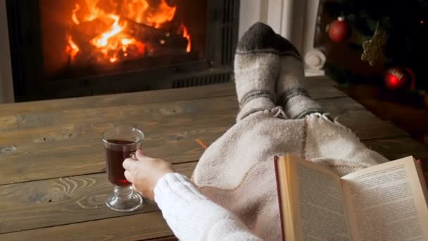 Slow motion footage of young woman reading book and drinking tea by the fireplace — Stock Video