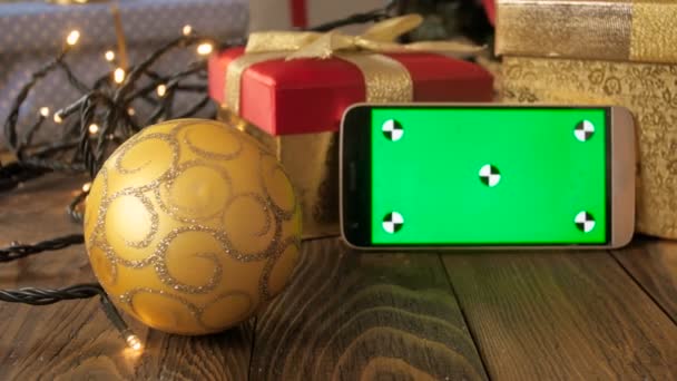 Крупный план съемки мобильного телефона с зеленым экраном Chroma Key на рождественском столе рядом с безделушками и подарочными коробками — стоковое видео