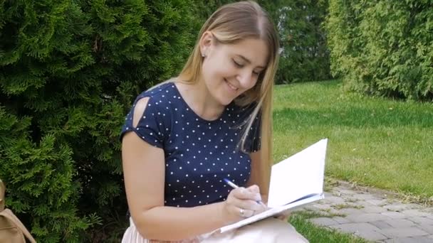 . Повільний рух кадрів красивої молодої жінки, що сидить у парку на траві і пише щоденник — стокове відео
