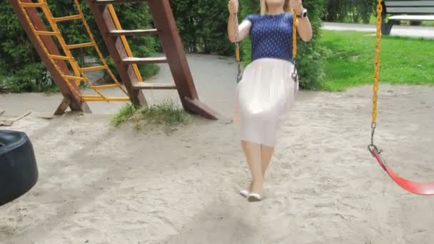 4K видео счастливой улыбающейся женщины в юбке, веселящейся на качелях в парке — стоковое видео