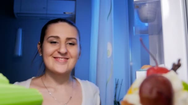 4 k πλάνα της νεαρής γυναίκας χαμογελαστά ψάχνει μέσα από το ψυγείο τη νύχτα και τη λήψη από σέλινο — Αρχείο Βίντεο