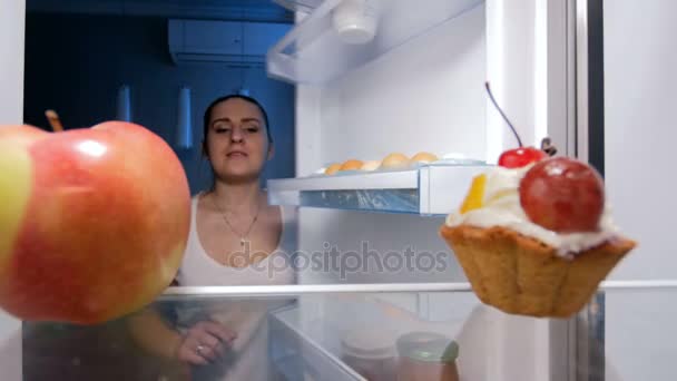4k 的镜头, 饥饿的年轻女子采取红苹果, 而不是从冰箱在晚上的蛋糕。节食的概念 — 图库视频影像