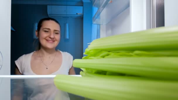 4k imagens de feliz mulher sorrindo à procura de comida na geladeira e mordendo aipo — Vídeo de Stock