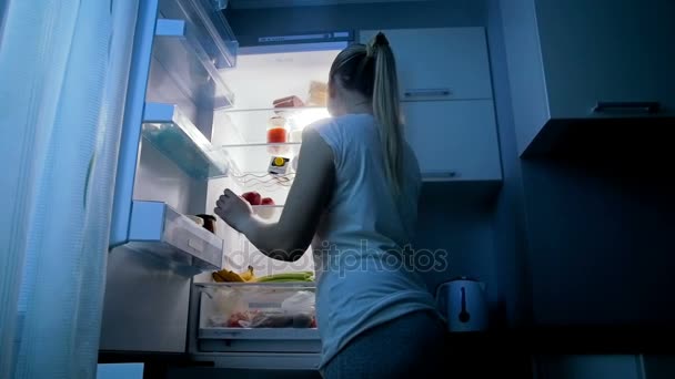Медленное видео, где молодая женщина забирает еду из холодильника ночью — стоковое видео