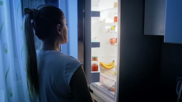 Slow motion video of young woman on diet looking inside of refrigerator at night — Stock Video