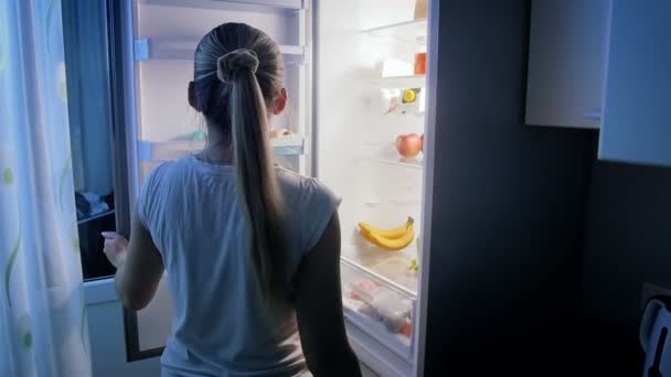 Slow motion video of young woman looking for food in refrigerator at night — Stock Video