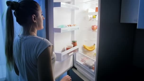 Ung kvinna i pajams öppnar kylskåpet på natten och letar efter mat — Stockvideo