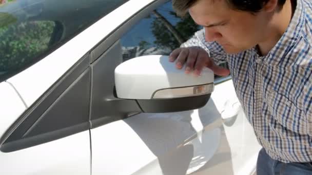 4k відео молодого чоловіка, який намагається видалити подряпину фарби на дзеркалі автомобіля — стокове відео