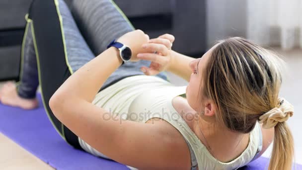 4 k video van jonge vrouw liggend op fitness mat met behulp van slimme horloge voor het meten van haar lichaamsparameters alvorens buik crunches te doen — Stockvideo